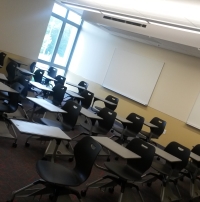 Photograph of a classroom on a sunny day. The classroom is at an angle, with disorderly rows of chair-desks, yellow walls, and a whiteboard. The classroom is empty of people.